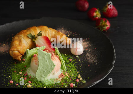 Fetta di strudel di mele con una pallina di gelato alla vaniglia su una piastra nera, su di un legno nero tabella, decorata con meringa e fragole. close-up Foto Stock