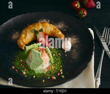 Fetta di strudel di mele con una pallina di gelato alla vaniglia su una piastra nera, su di un legno nero tabella, decorata con meringa e fragole. close-up Foto Stock