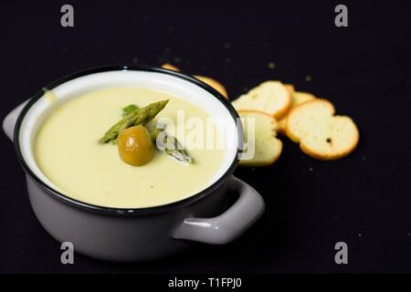 Cremosa zuppa di asparagi pieno di vitamine in grigio pot con olivi su sfondo nero Foto Stock