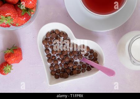 Le sfere di cacao cereali nel cuore bianco ciotola sagomata come parte di una completa prima colazione a buffet che include le fragole, il latte e la coppa di frutta rossa del tè. Parte superiore verso il basso Foto Stock