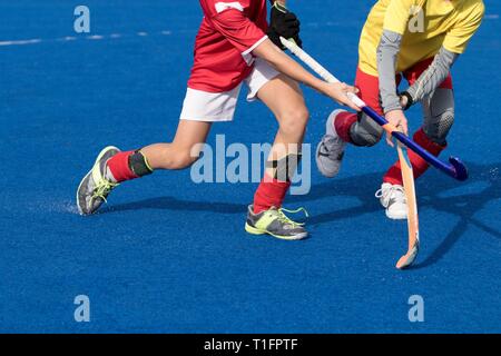 Campo di giovani giocatori di hockey su blu a base d'acqua turf. Azione intensa tra utente malintenzionato in rosso uniforme e difensore in giallo, indossando grigio sotto termica Foto Stock