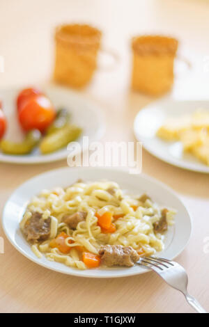 Pasta con le fette di carne e carote. Marinata di pomodori e cetrioli. Formaggio a fette in una piastra. Due tazze sul tavolo. Foto Stock