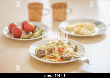 Pasta con le fette di carne e carote. Marinata di pomodori e cetrioli. Formaggio a fette in una piastra. Due tazze sul tavolo. Foto Stock