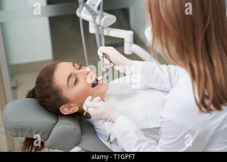 Felice paziente sdraiato sulla poltrona del dentista in odontoiatria clinica. Medico professionale tenendo il restauro degli strumenti e il trattamento di denti del client. Stomatologist indossando bianco uniforme e guanti. Foto Stock