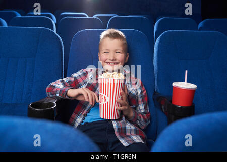 Divertente, gioiosa kid guardando film o cartoon commedia in cinema. Allegro Ragazzo che sorride, ridere, azienda big popcorn benna, seduta in blu scuro e confortevoli sedie. Foto Stock