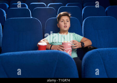 Ragazzo seduto in movie theater in confortevoli blu scuro sedia. Bel ragazzo holding benna popcorn, rosso bicchiere di carta con la bevanda frizzante. Concetto di intrattenimento e di svago. Foto Stock