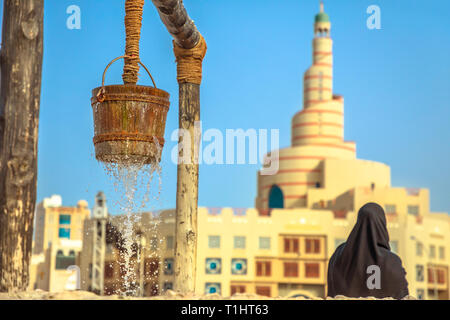 Primo piano di un flusso di acqua alla vecchia fontana al Souq Waqif nel centro di Doha con la donna dietro con vestito abaya e Moschea di Doha con minareto su sfocato Foto Stock