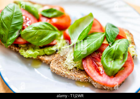 Primo piano di due strati di pezzi di tutto il grano germogliato di grano tostato pane sulla piastra con red le fette di pomodoro verde foglie di basilico e di avocado bruschetta su Foto Stock