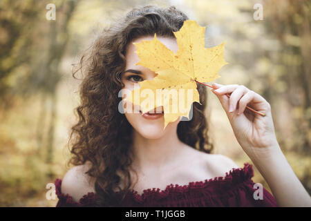 Bellissima ragazza con ricci capelli scuri in un marrone rossiccio top in un parco holding maple leaf davanti al suo volto e sorridente. Concetto di autunno. Foto Stock