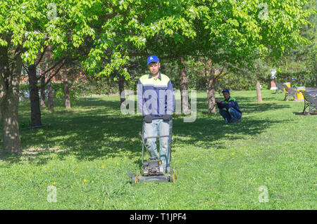 Mosca, Russia - 12 Maggio 2018: uomo taglio erba con tosaerba. Un altro uomo è in appoggio squatting Foto Stock