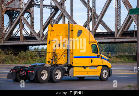 Di colore giallo brillante a lungo raggio professionale commerciale big rig semi carrello trattore in marcia su strada sotto il traliccio vecchio ponte ferroviario a magazzino per il pi Foto Stock