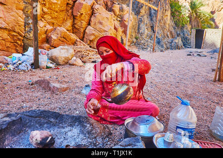 DAHAB, Egitto - 16 dicembre 2017: Il beduino ospitale donna in abito tradizionale e niqab tratta i turisti con il tè caldo, bollito su carbone di legna ho Foto Stock