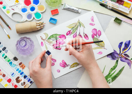Pittore tenendo un pennello in mano. Disegno ad acquerello - rosa fiori di ciliegio - e attrezzature artistico sulla scrivania. Vista dall'alto. Pittore disegno al lavoro Foto Stock