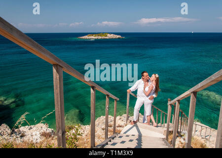 Vista romantica di coppia felice in bianche vesti coccola e in piedi sul litorale roccioso. Il pittoresco paesaggio marino di mare azzurro e la piccola isola. Concetto di ho Foto Stock