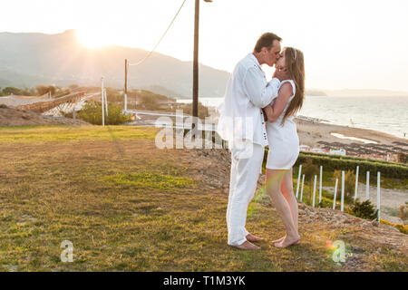 Vista romantica del giovane in bianche vesti quando essi kiss e stare a piedi nudi sull'erba verde collina. Lo splendido paesaggio di mare e di sole sopra mountai Foto Stock