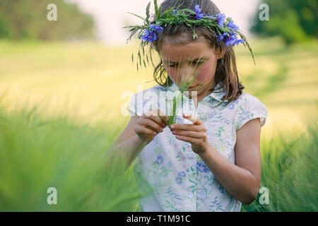 Curiosa la ragazza con i fiori nei capelli esaminando Grano verde levetta in campo rurale Foto Stock
