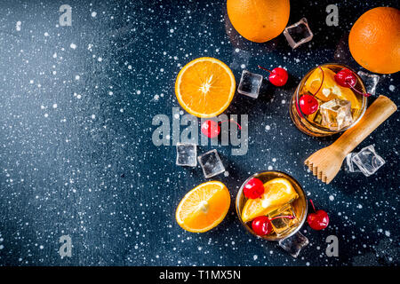 Classico cocktail di alcool, whiskey con cubetti di ghiaccio, fettine di arancia e cocktail ciliege rosse. In due bicchieri, su un blu scuro tabella di cemento, Foto Stock