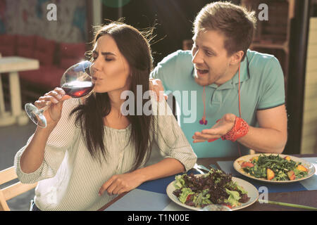 La risoluta ragazza dai capelli scuri bere il vino dal vetro mentre il suo fidanzato scuotendo il suo Foto Stock