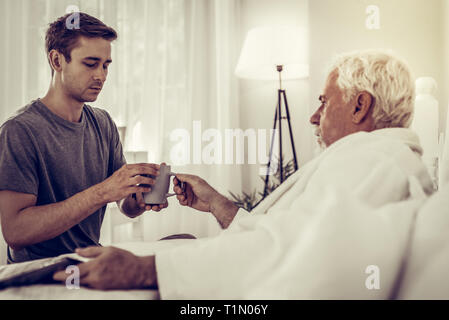 Focalizzata l uomo che passa la coppa al padre malato a letto Foto Stock