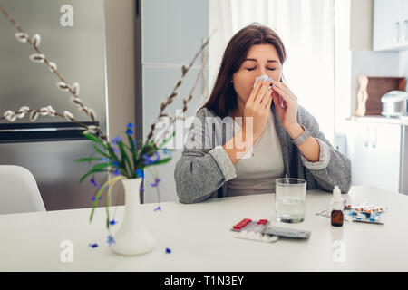 Allergia a molla. Giovane donna starnuti perché di fiori circondata con le pillole su cucina a casa. Allergia stagionali concetto. Foto Stock