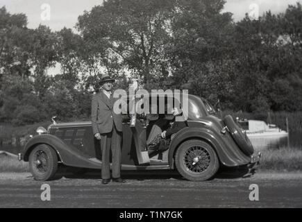 1930s, storico motoring in questa epoca, un gentiluomo in un vestito e indossando un cappello apre la porta sul retro della sua autovettura sportiva per la sua femmina passeggero come si fermano per una pausa da un bordo del fiume, Inghilterra, Regno Unito. Foto Stock