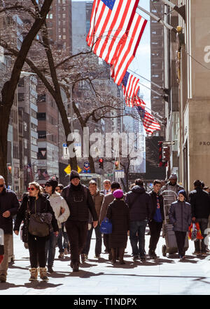 I pedoni a camminare lungo la Quinta Avenue, il centro di Manhattan, con bandierine americane volare al di sopra su una estesa pennoni. La città di New York, Marzo 2018 Foto Stock