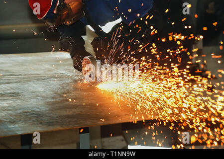 Un lavoratore in guanti e tuta taglia un foglio di metallo con un angolo-macchina rettificatrice. La formazione di scintille volare dal disco. Background Industriale con spazio per la copia Foto Stock