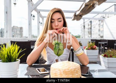 Ragazza di bere cocktail bar estivo sulla veranda del ristorante. Capelli lunghi, vetro di calce bere, cannuccia. Abito bianco sottile e conciate di figura Foto Stock