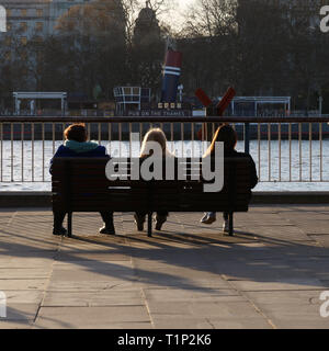 Tre persone sedute su una panca sulla banca del sud con una vista sul fiume Tamigi Londra, Regno Unito, e godetevi il sole del pomeriggio in un caldo giorno verso la fine di marzo. Foto Stock