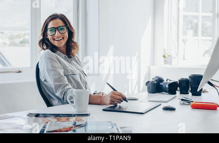 Fotografo professionista seduto alla sua scrivania in ufficio che guarda lontano e sorridente. Donna in ufficio digitale con tavoletta grafica e disegno penna. Foto Stock