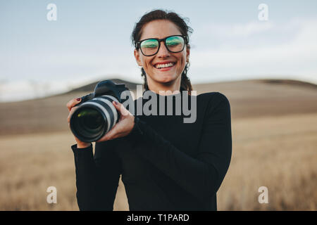 Fotografo positivo godendo foto riprese all'aperto. Giovane donna in casuale in possesso di una reflex digitale e sorridente. Foto Stock