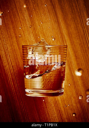 Bicchiere di vetro che cade con un cubetto di ghiaccio e acqua su di un legno Sfondo della granella Foto Stock