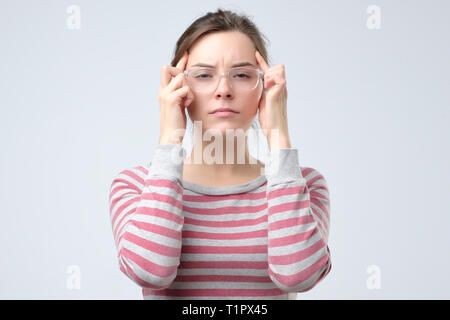 La donna mantiene entrambe le dita indice su templi, soffre di emicrania, indossa occhiali tondi, essendo cupa. Lei sta cercando di fare una scelta giusta o generi Foto Stock