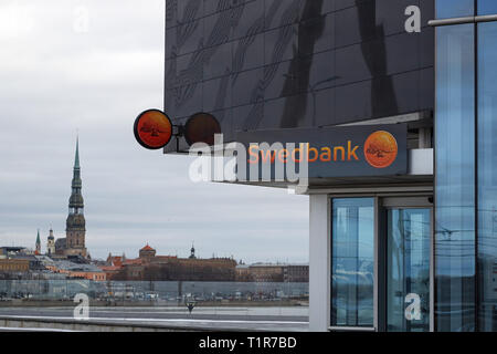02 marzo 2019, la Lettonia, Riga: una filiale della banca svedese Swedbank può essere visto al piede di un vetro edificio per uffici. Sullo sfondo si può vedere il panorama della città vecchia di Riga con la Petrikirche (M). Swedbank è la più grande banca in Svezia e gli Stati baltici, secondo le proprie cifre. Essa aveva dichiarato che aveva riportato un totale di 3774 le operazioni sospette alle autorità negli Stati del Baltico nel 2017 e 2018. In futuro si intende intensificare la lotta contro la criminalità dei colletti bianchi mediante la costituzione di un gruppo speciale all'interno della banca. Foto: Alexander Welscher/dpa Foto Stock