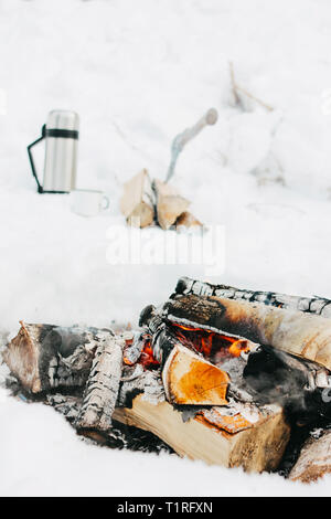 Legna da ardere con carboni nel fuoco nella neve sullo sfondo dei thermos e ascia di guerra. Concetto di viaggio Foto Stock