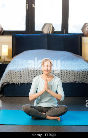 Tranquilla donna matura in vestiti comodi facendo yoga nel suo elegante appartamento Foto Stock