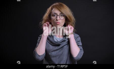 Closeup ritratto di mezza età stravagante redhead femmina in bicchieri facendo un divertente di espressione del viso mentre guardando dritto in telecamera Foto Stock