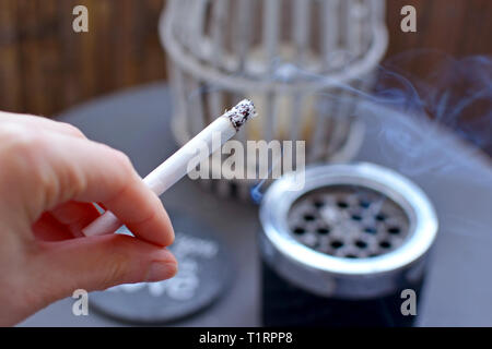 In prossimità di una donna di mano che tiene un acceso il fumo di sigaretta sfocata con posacenere sul tavolo in background Foto Stock