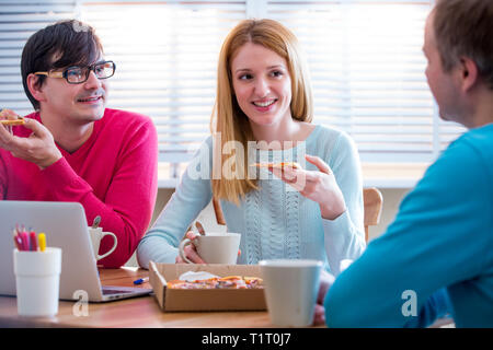 Happy business persone aventi una riunione e mangiare la pizza in ufficio. Tre giovani colleghi allegro a pranzo insieme al lavoro. Il team Creative intorno Foto Stock