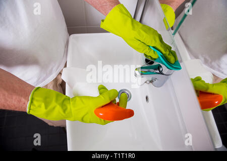 Uomo in gomma guanti protettivi per la pulizia e la lucidatura lavello e rubinetto. Superficie lucida del rubinetto di acqua, mani con spugna e bottiglia di agente di pulizia. Foto Stock