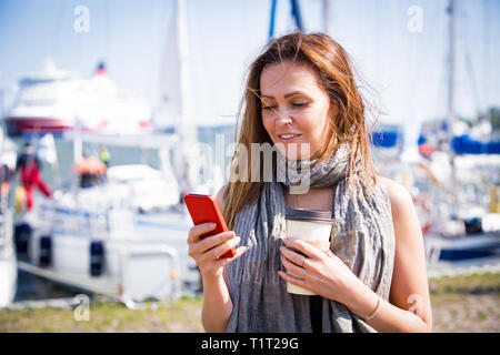 Ritratto di sorridente donna elegante con smart phone in mano a piedi lungo la baia mare. Barche a vela nel porto di mare. Texting, surf, bere caffè. Foto Stock