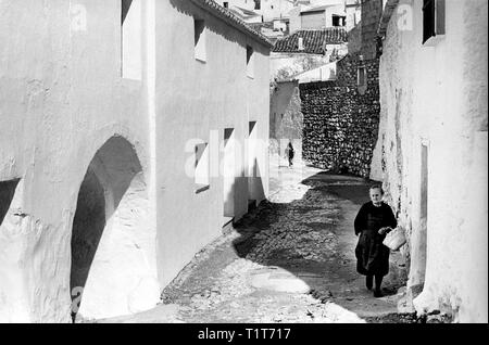 Mijas, vicino a Malaga, Spagna 1968 - una vecchia donna vestita di nero tradizionale passeggiate al mercato con un sacco di bambini giocare puggyback in strada bey Foto Stock