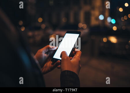 White schermo vuoto del telefono intelligente in mani femminili. Giovane donna la digitazione sul dispositivo mobile mentre si sta in piedi sul street nella città di notte, luci incandescente su ba Foto Stock