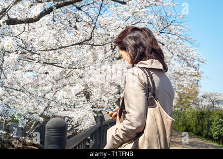 Una donna con uno smartphone che indossa una maschera facciale e un cappotto beige. Periodo primaverile durante Covid-19. Foto Stock