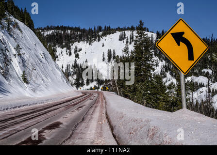 Curva a sinistra segno: Gli automobilisti sono avvisati di un forte giro precedendo su una montagna innevata strada in Montana. Foto Stock