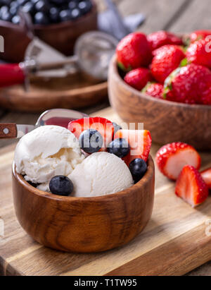 Gelato alla vaniglia con mirtilli freschi e le fragole in un recipiente di legno con ciotole di frutti di bosco in background Foto Stock