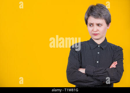 Stupito introverso giovane donna, che indossa la maglia nera, mantiene con le braccia incrociate guardando stupito da parte isolate su parete gialla con copia spazio. Confondere hip Foto Stock