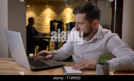 Closeup shoot di adulto imprenditore caucasico digitazione sul laptop e prendere appunti in ambienti chiusi in ufficio Foto Stock