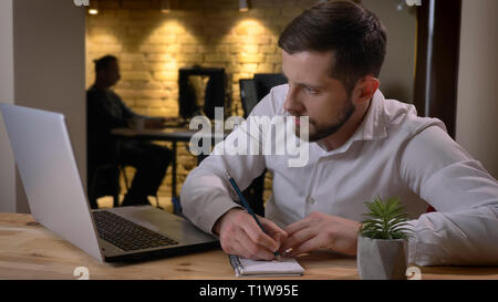 Closeup shoot di adulto imprenditore caucasico digitazione sul laptop e prendere appunti in ambienti chiusi in ufficio Foto Stock