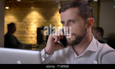 Closeup shoot di adulto imprenditore caucasico digitazione sul laptop e avere una formale chiamata telefonica in ambienti chiusi in ufficio Foto Stock